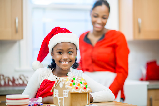 A mother and daughter are decorating a gingerbread house at home in their kitchen. They are both wearing red and the little girl is wearing a Santa hat in the spirit of the Christmas holidays.