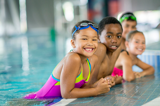 A multi-ethnic group of elementary children are taking swimming lessons together at the pool. They are smiling and looking at the camera.