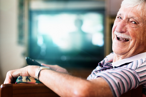 Laughing 90-year-old old man is happily relaxing by watching television, smiling over his shoulder at camera as he holds the remote control, in charge of everyone's viewing! Copy space on the defocused TV screen.