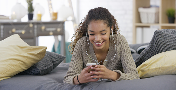 A mixed race teenage girl is enjoying time on her smart phone in her bedroom. Here, she listens to music with headphones.