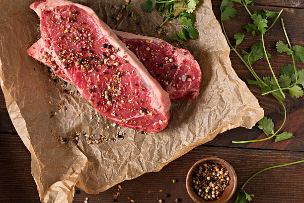 Two Raw New York Steaks An overhead extreme close up horizontal photograph of two New York strip stakes on brown butcher paper, seasoned with  flake salt and pepper and garnished with some fresh green cilantro. raw steak beef meat stock pictures, royalty-free photos & images