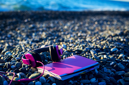 Pink headphones lying at paper notebook on sea beach. Closeup. Working concept. Freelancer. Music listening, Travel.