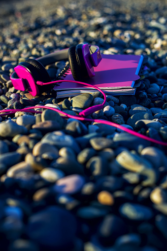 Pink headphones lying at paper notebook on sea beach. Closeup. Working concept. Freelancer. Music listening, Travel. Vertical.