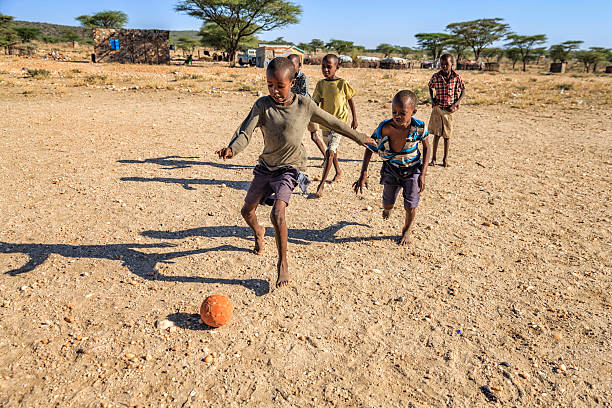 bambini africani a piedi nudi che giocano a calcio nel villaggio, africa orientale - povertà africa foto e immagini stock