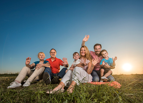 Smiling extended family relaxing in grass at sunset while waving and looking at the camera. Copy space.