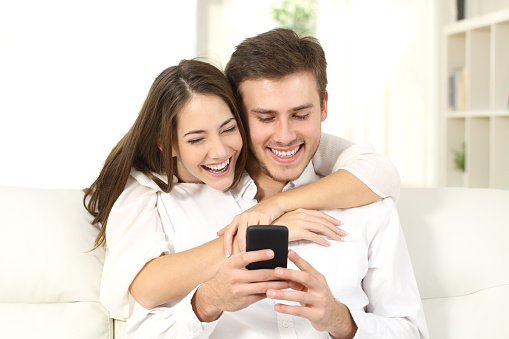 Funny couple or marriage sharing a smart phone to watch media content sitting on a couch at home