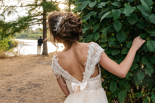 Bride peeking out from behind a tree. groom standing at a distance. Wedding photo shoot, rear view.