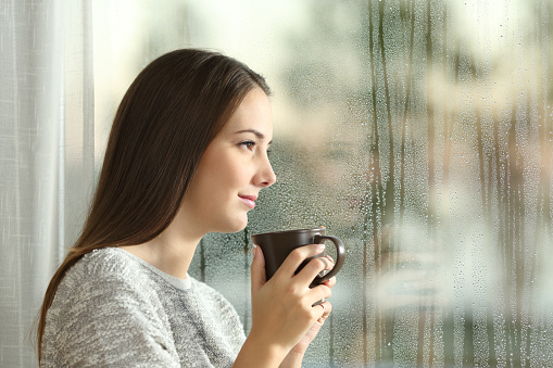 Side view portrait of a pensive woman looking away through a wet window in a rainy day at home