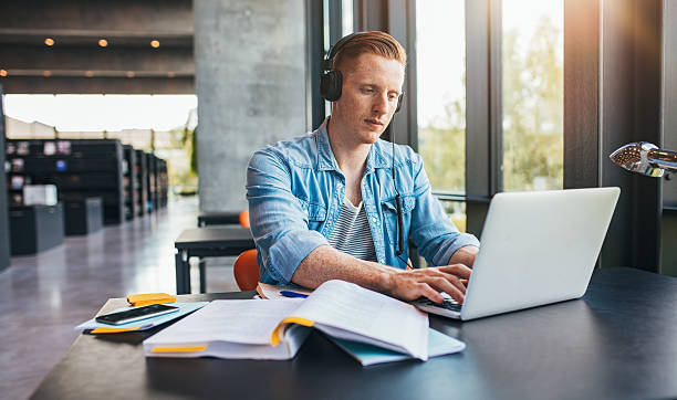 Handsome male student in a university library Handsome male student in a university library working on laptop. Young man studying on school assignment. jacob ammentorp lund stock pictures, royalty-free photos & images