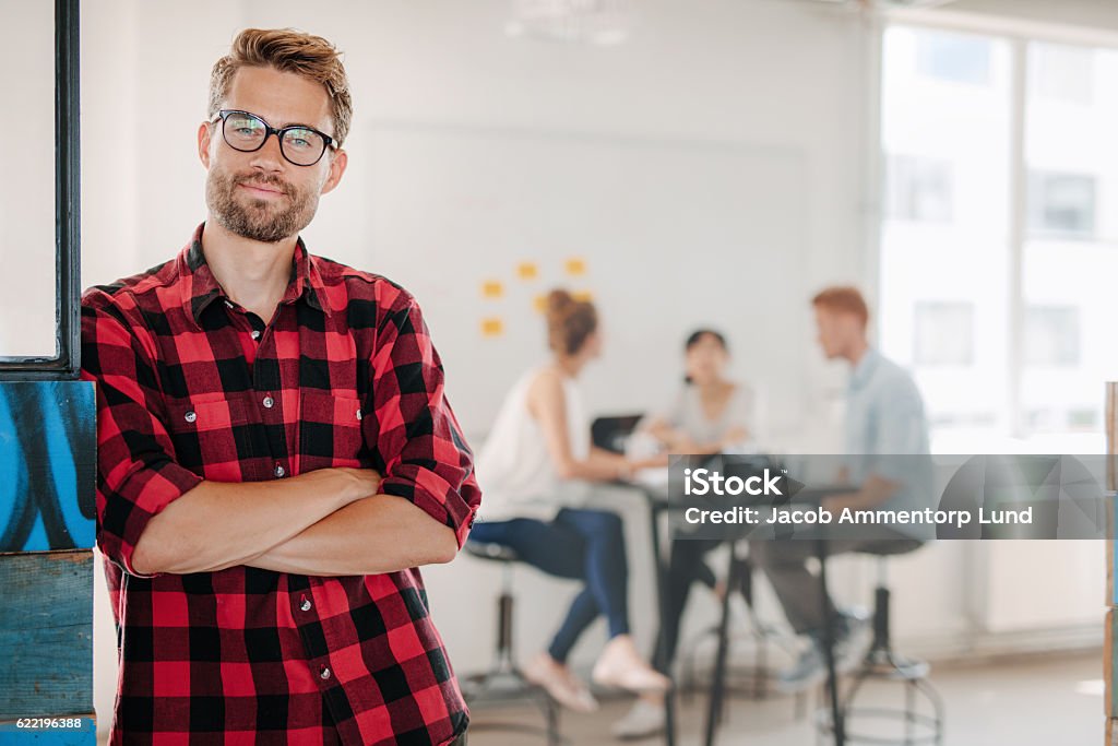 Relaxed business man in office with colleagues in background Portrait of relaxed young man standing in office with colleagues meeting in background. Young Men Stock Photo