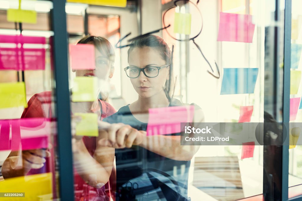 Two business women working together on wall glass Two business females working together on wall glass with post it stickers. Modern startup office Adhesive Note Stock Photo