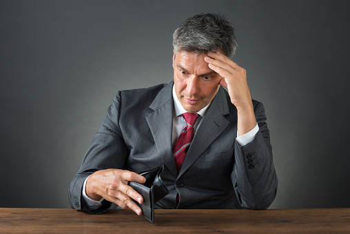 Shocked businessman with empty wallet sitting at desk against gray background