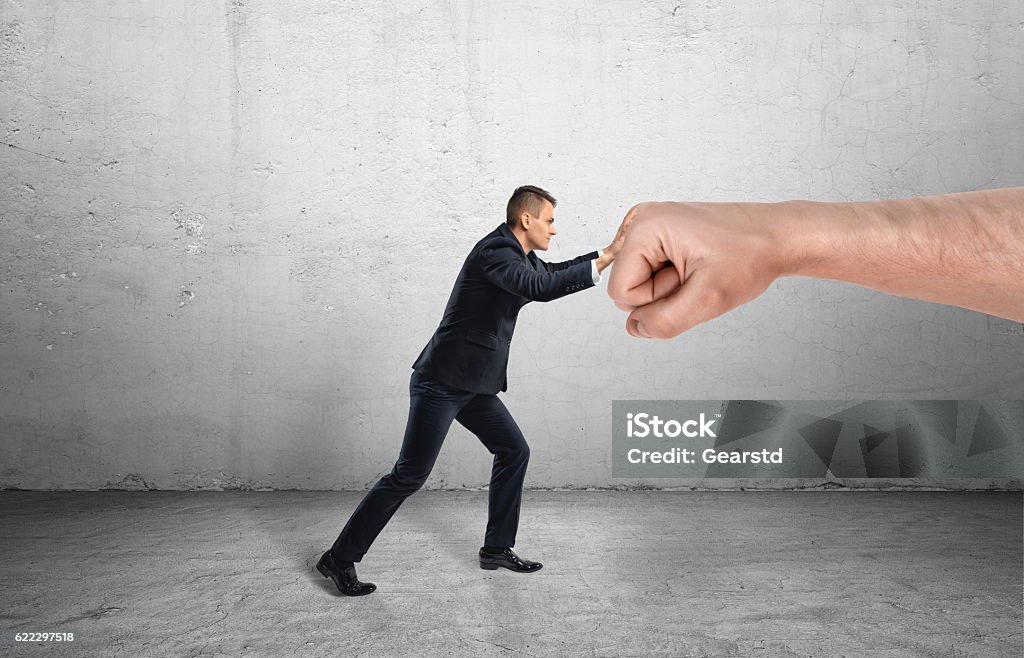 Businessman trying to resist huge male fist and move it Businessman trying to resist a huge male fist and move it away on a grey background. Containing market pressure. Being competitive. Protecting a small business. Small Stock Photo
