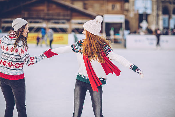 Friends having so much fun while ice skating. Wearing warm clothing. City is decorated with christmas lights.