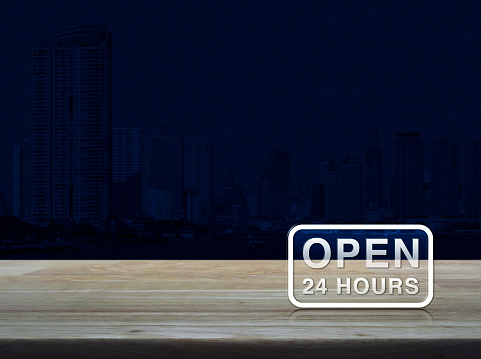 Open 24 hours icon on wooden table over modern office city tower blue tone background