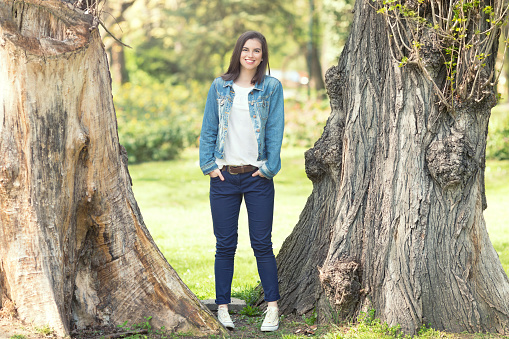 Beautiful young woman looking at camera and smiling while between trees in a park on a sunny spring day. She is standing with her hands in pockets wearing denim jacket and blue pants. Copy space