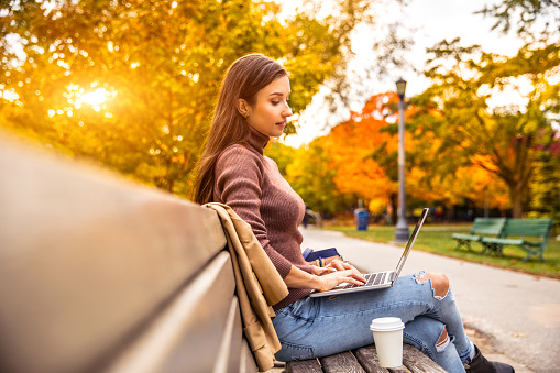 Woman working outdoor sitting on a park bench in fall season.