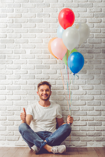 Handsome guy in casual clothes is holding balloons, showing Ok sign and smiling, sitting on the floor, on white brick wall background