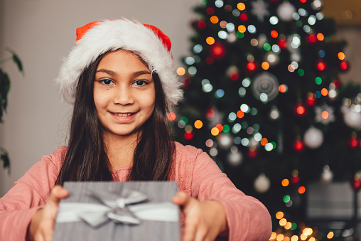 Girl with santa hat holding christmas gift and sitting by the christmas tree