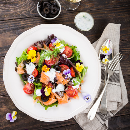 Fresh salad with smoked salmon, black olives, cherry tomatoes and edible flowers on wooden background. Top view.