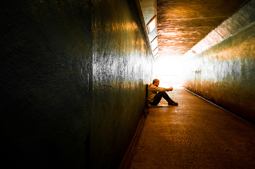 Wide angle image of a young man of caucasian ethnicity sitting on the floor of a subway tunnel and holding out a paper cup to beg for money. He is wearing jeans and a hoodie and has his head bowed in shame, despair and sadness. The tunnel is bathed in a golden, warm light. Horizontal colour image with copy space.