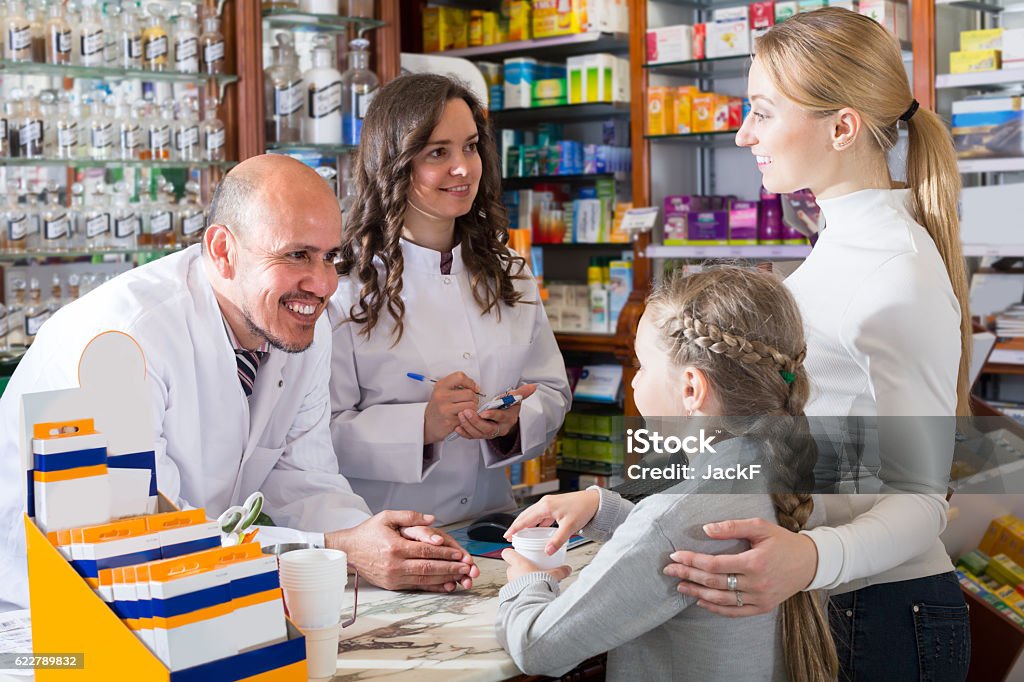 Two pharmacists and customers Two friendly smiling pharmacists wearing white coats standing next to cashier and consulting a customer with child. Selective focus Family Stock Photo