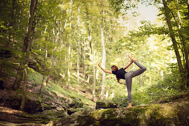 Beautiful middle aged woman practicing yoga in wood next to the river. She's wearing a sports clothing. She is standing barefoot on a rock and stretching