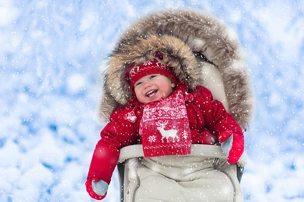 Happy laughing aby in stroller in winter park with snow Happy laughing baby in warm red down jacket and knitted Nordic hat and scarf on a walk in a snowy winter park sitting in warm stroller with sheepskin hood catching snow. Child in buggy with foot muff baby stroller winter stock pictures, royalty-free photos & images