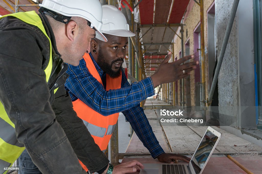 Two construction workers, an African american and a white Two construction workers, an African american and a white, wearing orange and yellow safety jackets and helmets among scaffolding on construction site Construction Site Stock Photo