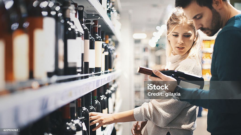 Couple buying some wine at a supermarket. Closeup side view of late 20's couple choosing some red wine at local supermarket. The guy is holding on of the bottles and they're both reading the label on the back. Big selection of unrecognizable red wines in front of them. Wine Stock Photo