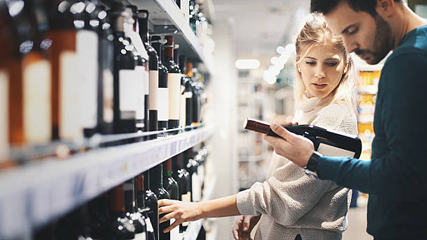 Couple buying some wine at a supermarket. Closeup side view of late 20's couple choosing some red wine at local supermarket. The guy is holding on of the bottles and they're both reading the label on the back. Big selection of unrecognizable red wines in front of them. alcohol shop stock pictures, royalty-free photos & images