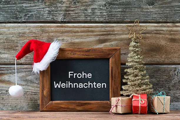 Merry Christmas decorations, chalkboard with german text Frohe Weihnachten means Merry Christmas and a red Santa hat, small christmas tree and gifts on a timber background