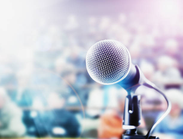 Brightly lit microphone in front of out-of-focus audience A vocal microphone is brightly lit in front of a defocussed audience on tiered seating who provide ample copy space. microphone stand photos stock pictures, royalty-free photos & images