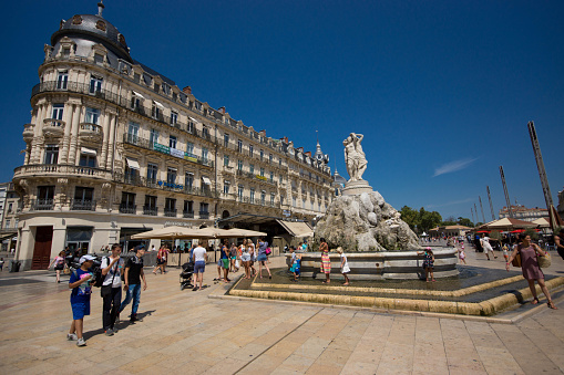 Montpellier, France - August 5, 2015: Plaza de la Comedia in Montpellier France with tourists walking across the plaza. The fountain of 3 graces (Fontaine des Trois Graces) is in the foreground.