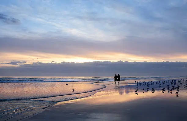 Photo of Senior couple holding hands enjoying time on beach at sunrise.