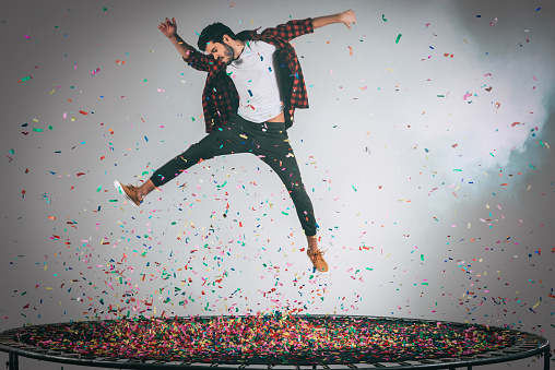 Mid-air shot of handsome young man jumping on trampoline with confetti all around him