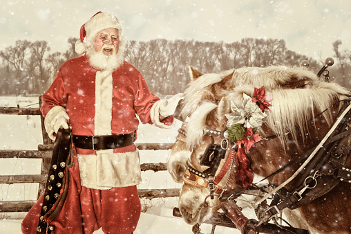 Santa in an outdoor setting talking to his team of horses.  He is holding a set of handbells in his right hand.  A light winter snow is falling.