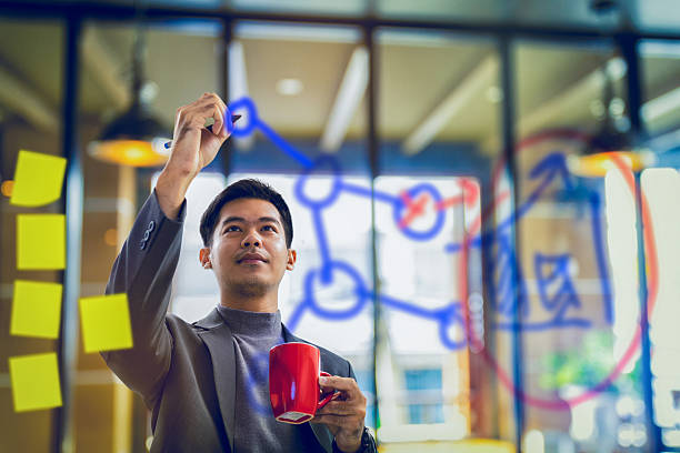 businessman holding coffee mug and writing data on transparent businessman holding coffee mug and writing data on transparent board mirror writing stock pictures, royalty-free photos & images