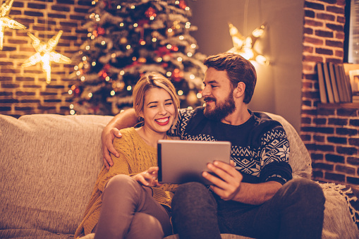 Couple sitting on sofa at home. Wearing Christmas sweaters. Together enjoying in the warmth of their home for Christmas. Using digital tablet.