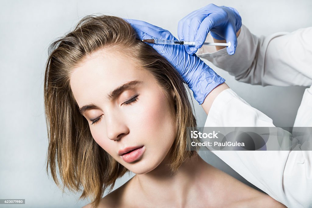 Doctor aesthetician makes head beauty injections to beautiful female patient Doctor aesthetician in blue medical gloves makes hyaluronic acid rejuvenation beauty injections in the back of the head of beautiful young female patient for hair growth and to prevent boldness. Adult Stock Photo