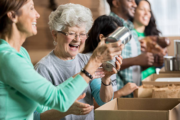 Happy female friends volunteer at charity food bank Mature and senior Caucasian friends pack canned goods into boxes during community food drive. Volunteers are working in the background. charitable foundation photos stock pictures, royalty-free photos & images