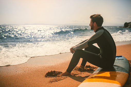 Portrait of a young surfer in a wetsuit, sitting on a surfboard and watching the waves