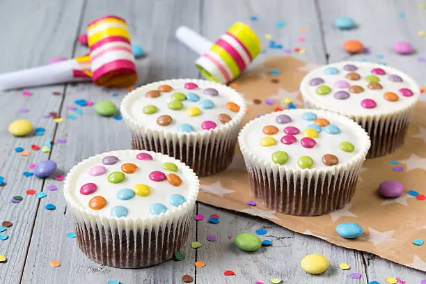 Cupcakes with white icing and colored smarties, party horns and confetti, wooden background