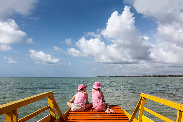 A DSLR Canon photo of two brazilian girls enjoying their vacations on a bright sunny summer morning in Porto de Pedras city, Alagoas, Northeastern Brazil. They are sitting on the prow of a small boat with their backs to the camera. The sea is flat and there are some white clouds in the blue sky.