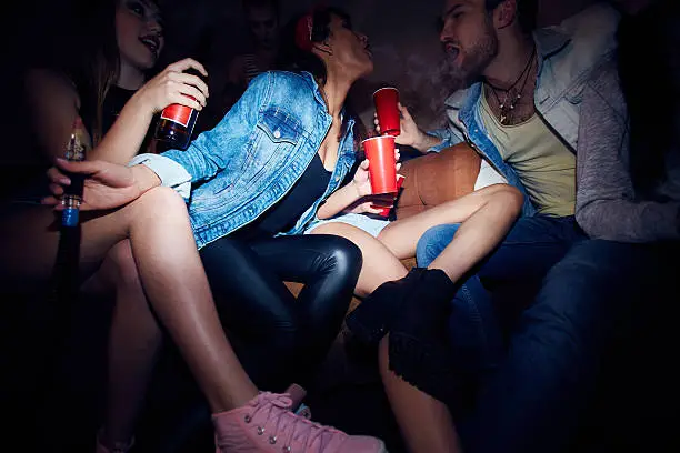 Sneak low angle shot of modern young party people, group of girls and man, sitting on sofa in chilling lounge of dark club, drinking beer and blowing vape smoke out to each others mouth