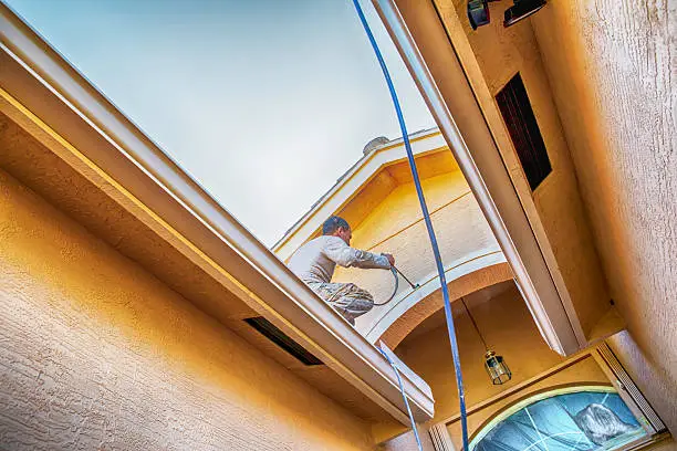 A hispanic house painter is standing on a roof while using an electric power spray painter to paint the outside of a home. He is painting the trim above the entryway. Taken with Canon 5D Mark IV.