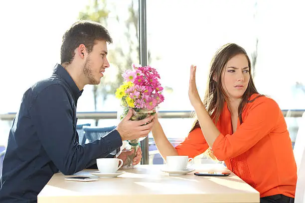 Man asking for forgiveness offering a bunch of flowers to his girlfriend in a coffee shop with an exterior background. Couple problems concept