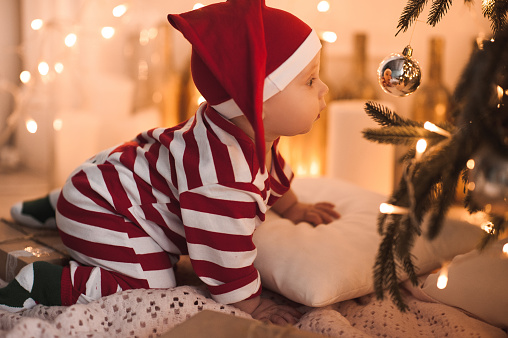 Baby girl 1 year old crawling under Christmas tree wearing body suit in red and white color. Looking at Christmas ball and see her reflection. Holiday season.