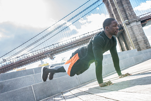 Young black man exercising outdoors. He is doing push-ups in the city. Bridge in the back.