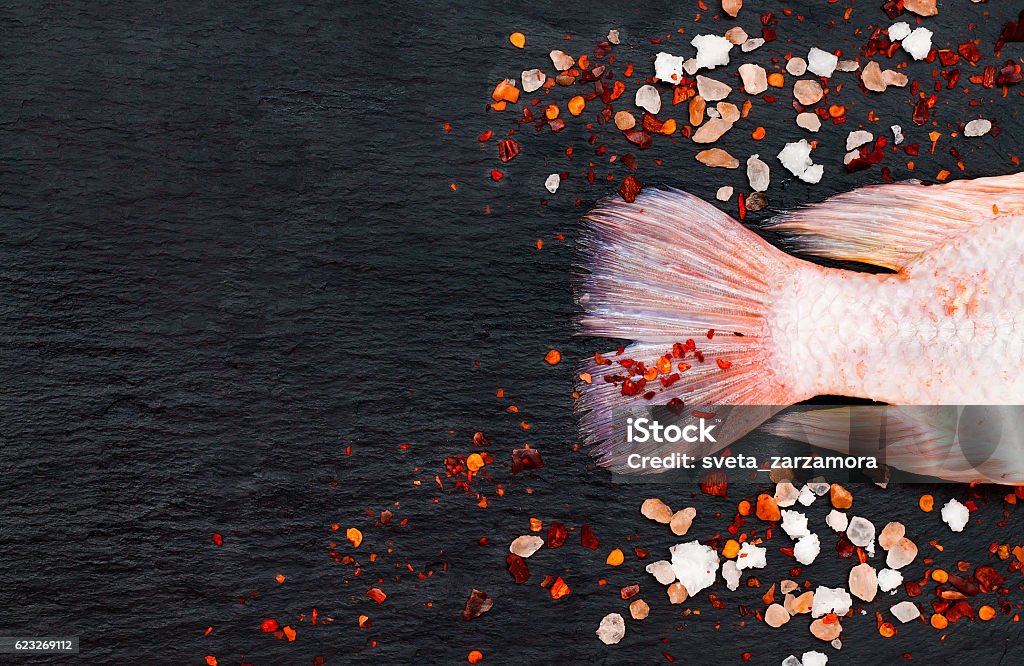 Tilapia fish on black board with spices. Culinary seafood background Tail of fresh raw red tilapia fish on black slate stone board with spices and pink himalayan salt. Culinary seafood background. Top view, copy space Above Stock Photo